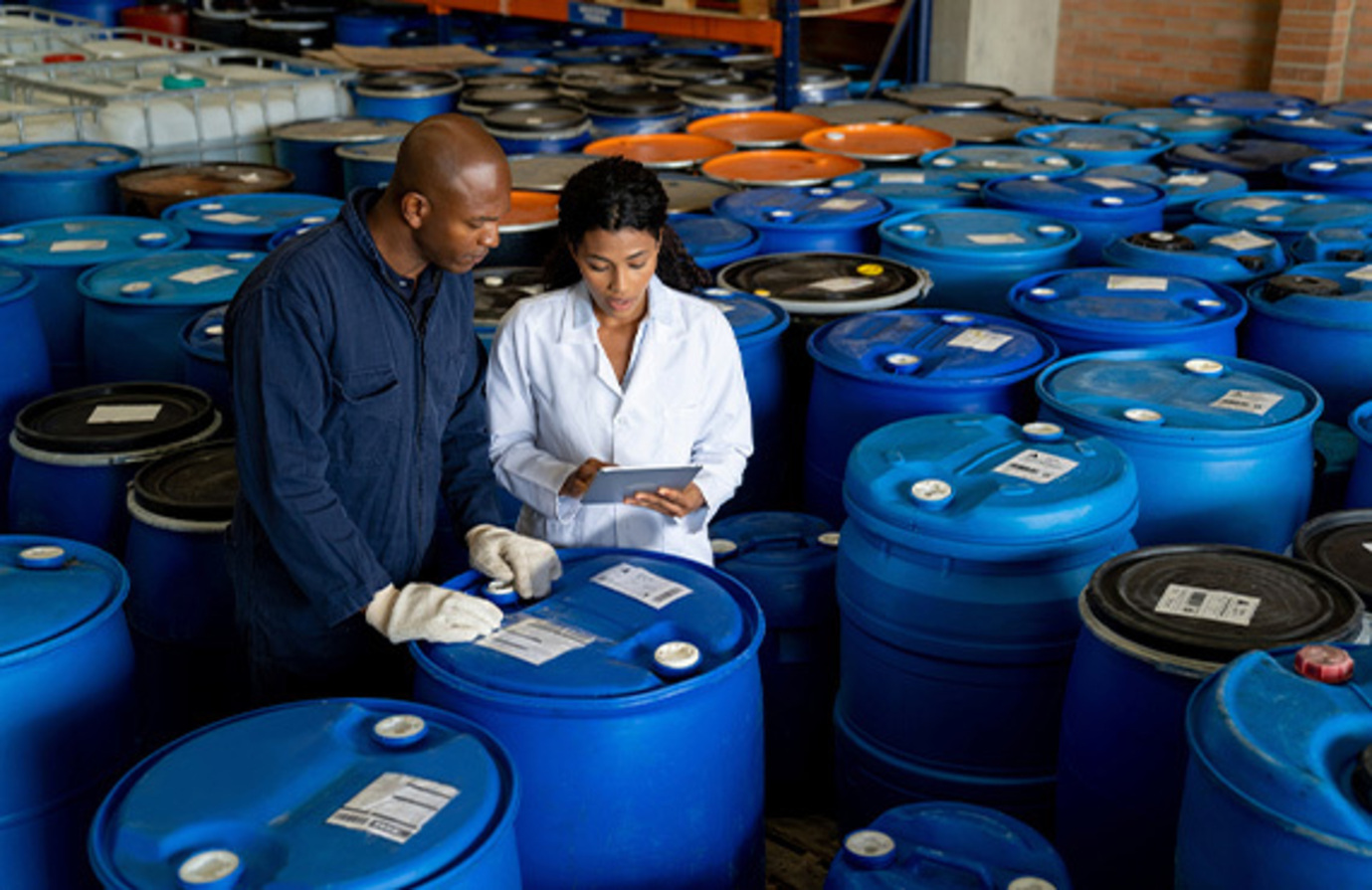 Two chemical plant workers surrounded by chemical barrels look at data on a tablet, highlighting the importance of protecting operations and automation with cybersecurity.