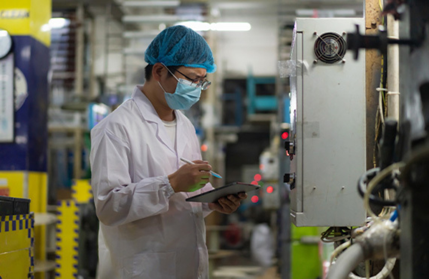 A man holds a tablet computer at a manufacturing facility, focusing on cybersecurity.