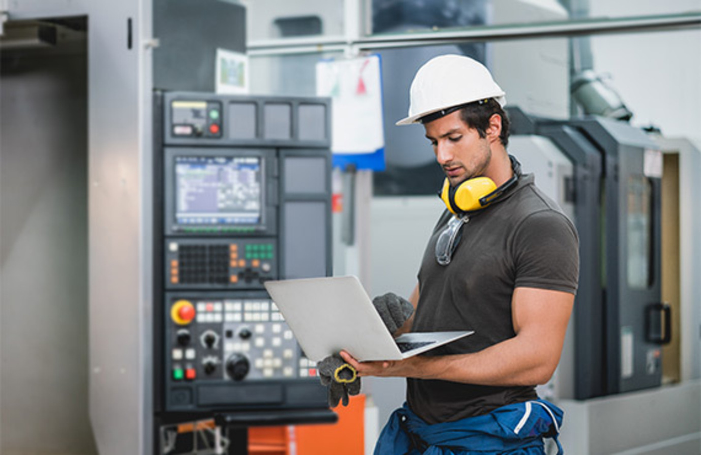 A man in a hard hat and safety glasses using a laptop in a factory for preventative maintenance.